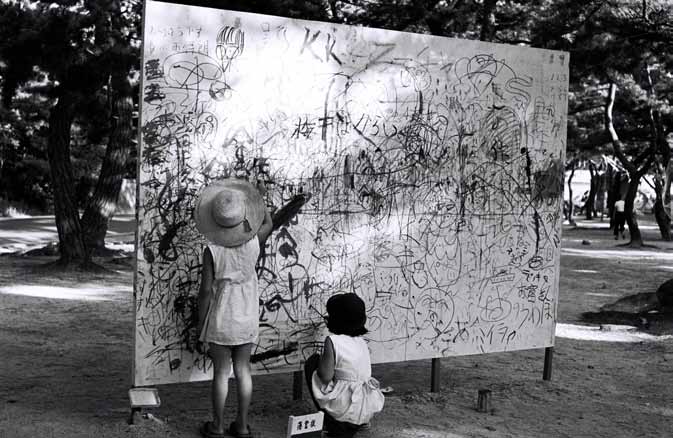 Children and other visitors draw on a large outdoor board covered with marks, watched by adults in Ashiya Park.