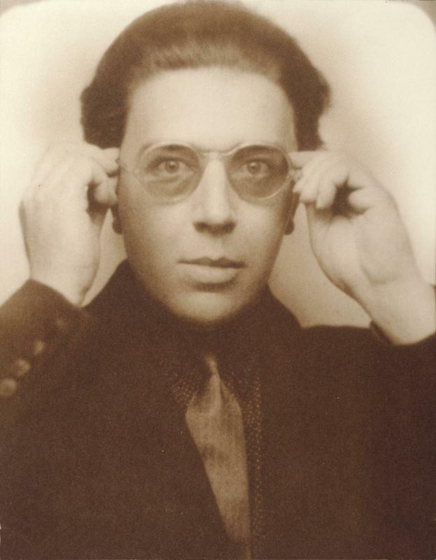 Sepia photobooth portrait of André Breton looking toward the camera with both hands raised to his round glasses.