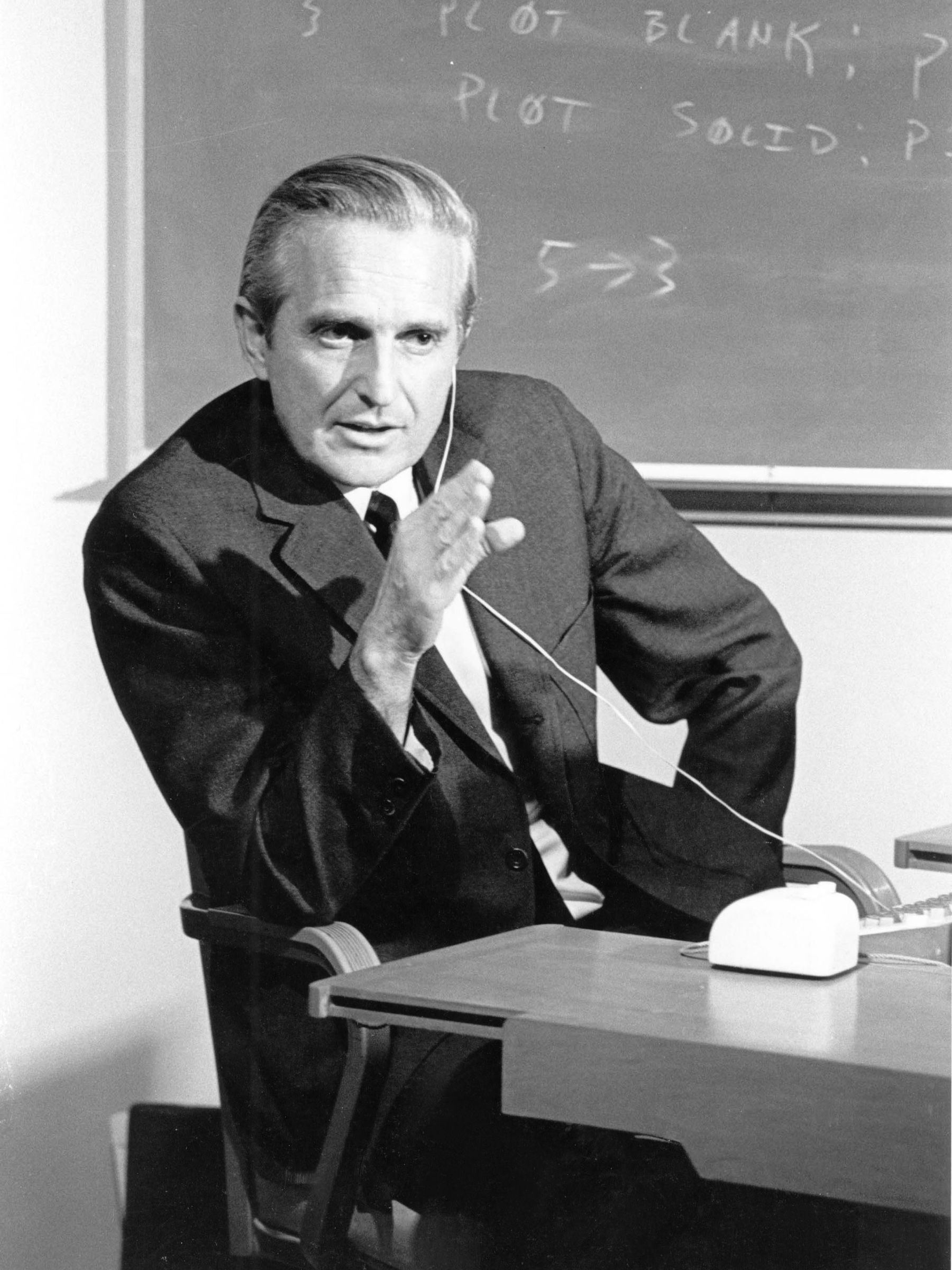 Douglas Engelbart gestures beside an early computer mouse, wearing an earpiece, with a blackboard behind him.