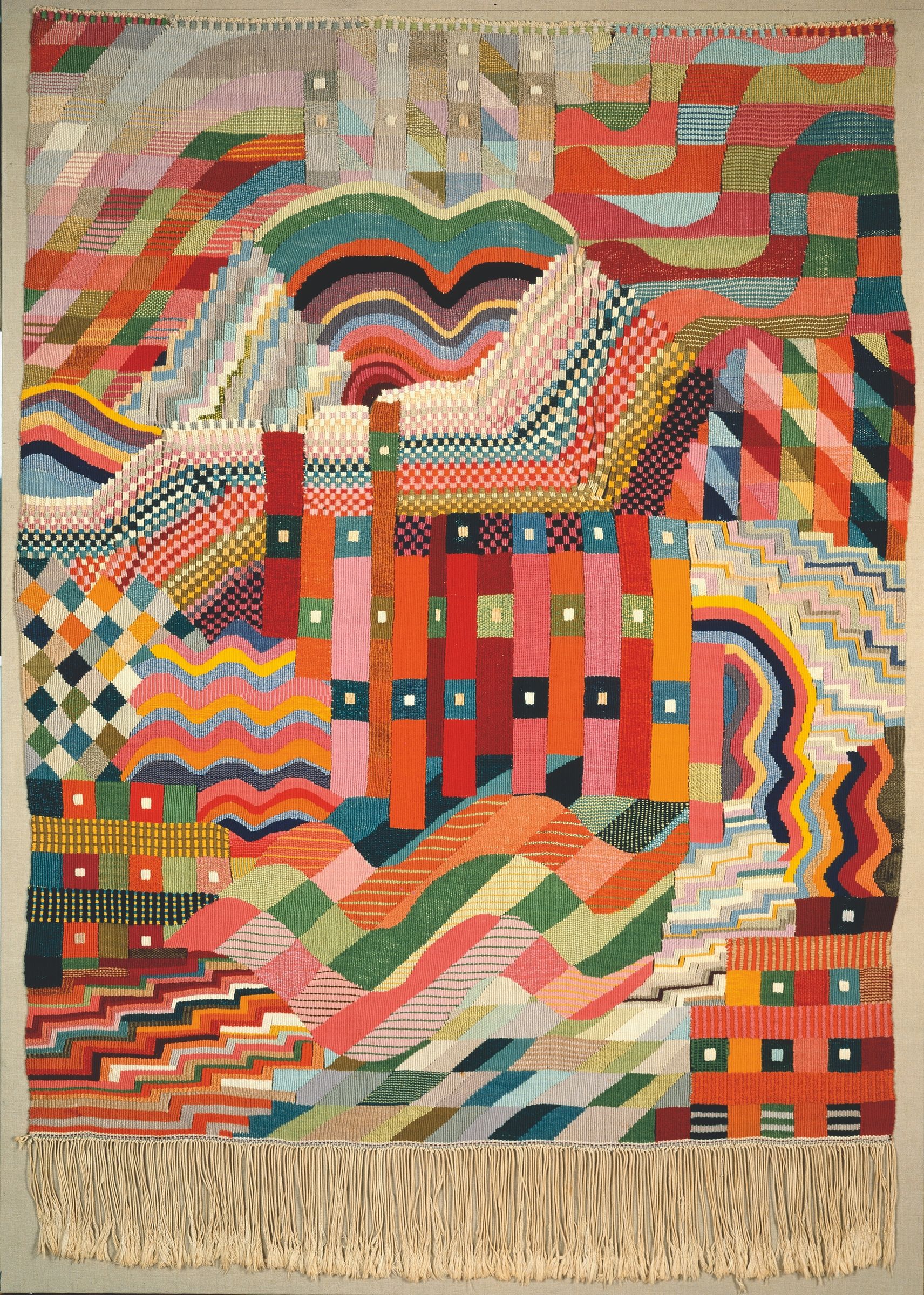 A full-length tapestry of vivid interlocking waves, stepped shapes, checkerboards and coloured vertical bands, with its fringe visible.