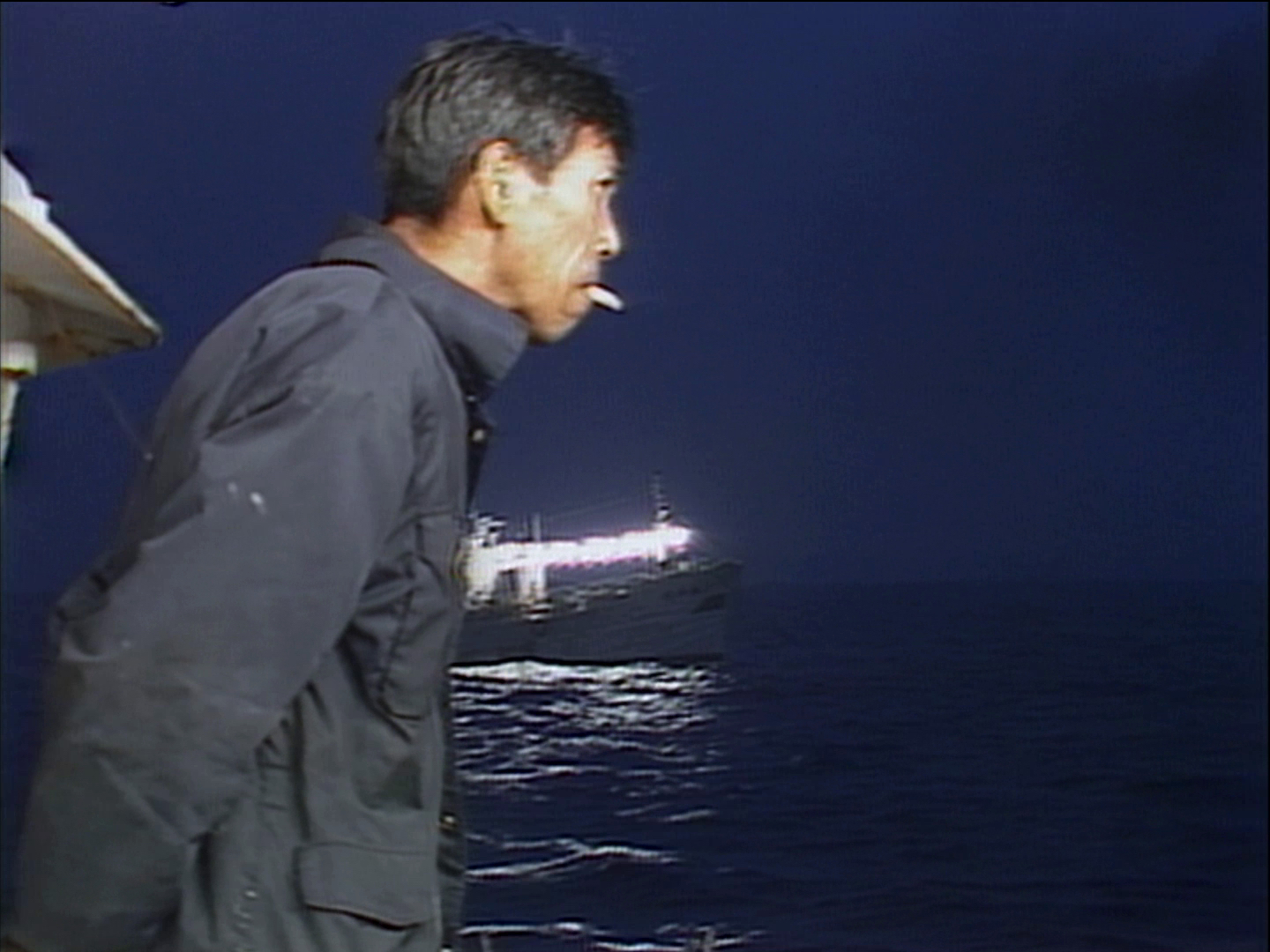 Video still of a man in a dark jacket beside water at night, with a lit boat and reflected light behind him.