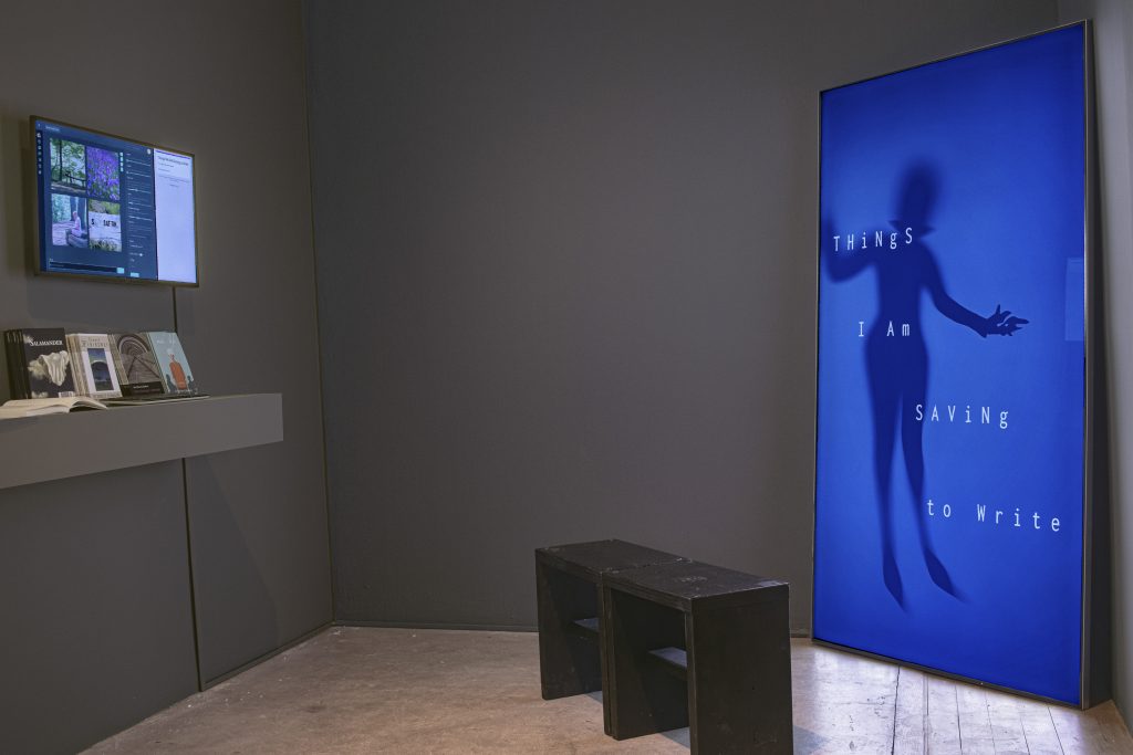 A tall cobalt-blue screen with a human silhouette and scattered words lights a gray gallery corner in Code Chronicles.