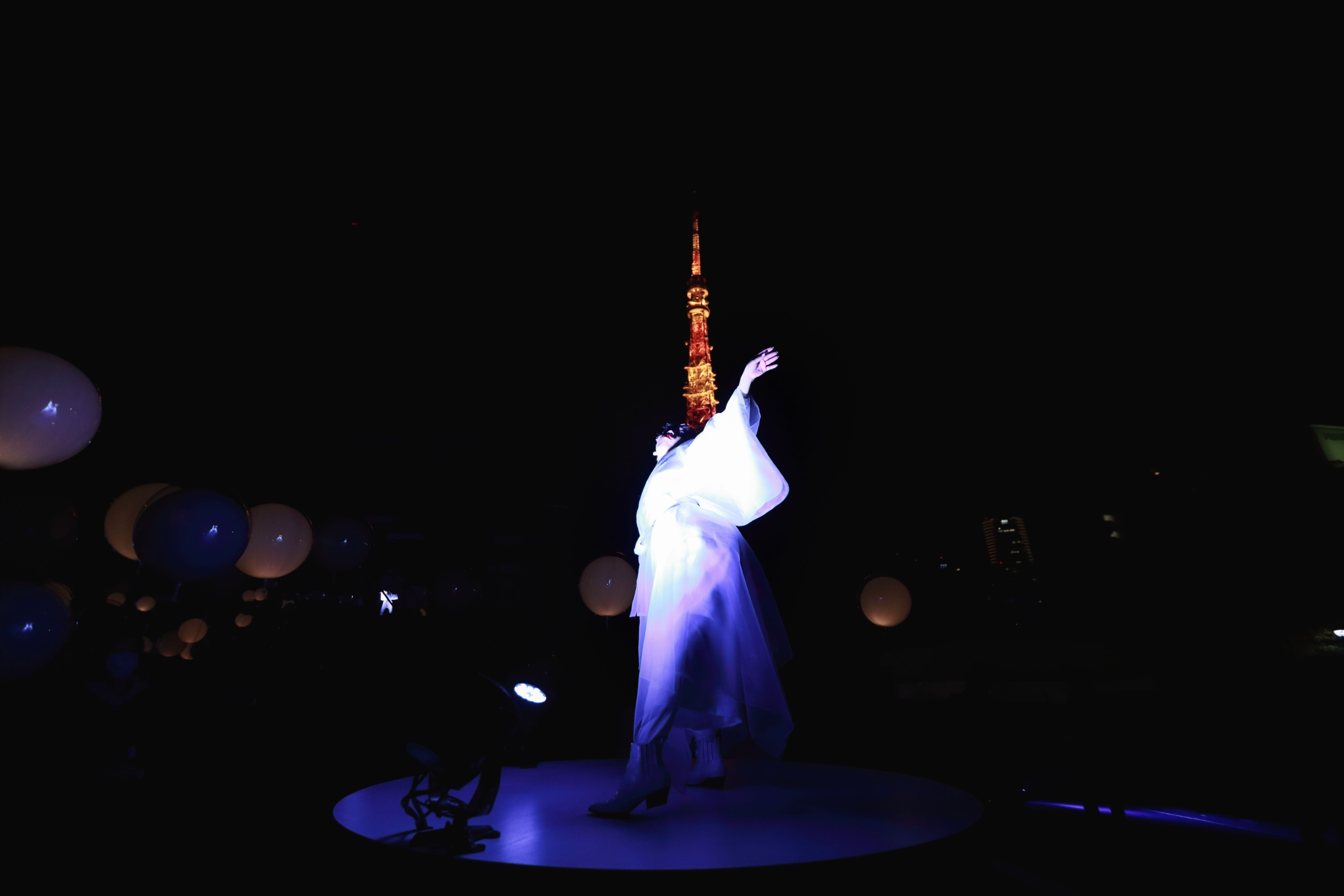 A dancer dressed in white is lit blue on a circular platform amid glowing balloon-like devices, with Tokyo Tower behind the night performance.