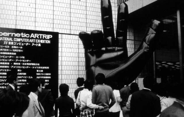 Passers-by outside the Sony Building in Ginza in October 1973, looking up at Shiro Takahashi's giant black inflatable hand beside the exhibition sign reading 'Cybernetic ARTRIP — International Computer Art Exhibition'.