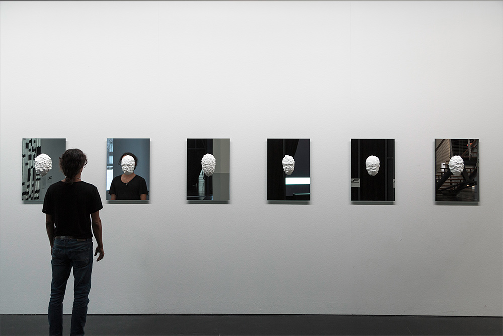 An installation view from Sterling Crispin's own documentation of Data-Masks at ZKM Karlsruhe, showing a row of the framed mask works on a gallery wall with a visitor in front of them.