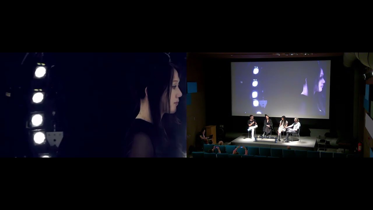 A conference video combines a remote participant with the seated panel on stage.