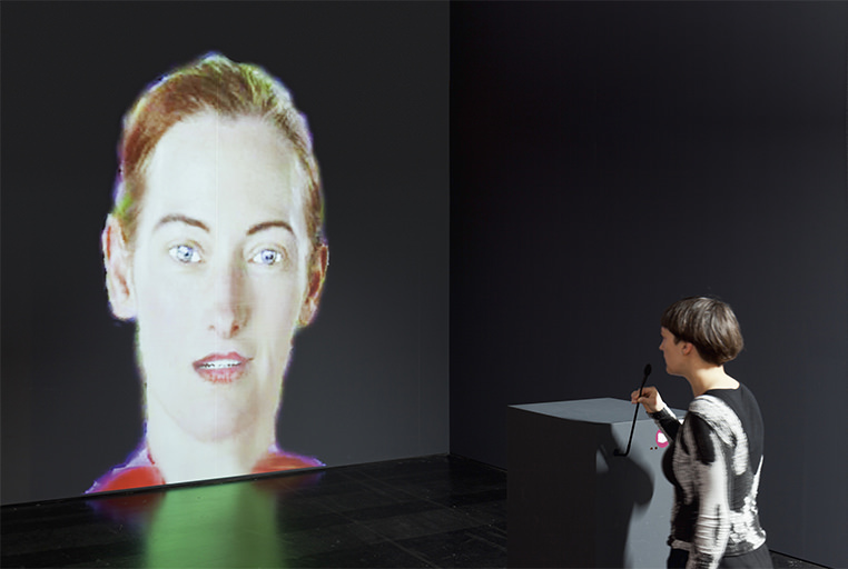An installation view of Lynn Hershman Leeson's DiNA, with a visitor speaking into a microphone at the podium while the projected agent answers.