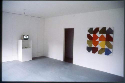 An installation view of Ernest Edmonds's 1985 one-person show at Exhibiting Space, London, with a monitor on a plinth showing the generative time-based work Fragments beside one of his systems paintings.
