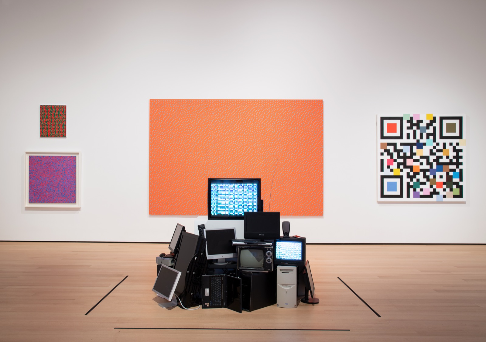 An installation view of the exhibition, not a single work: a gallery hung with a large orange monochrome, two small Op prints and a pixelated colour-block painting, with a heaped stack of obsolete televisions, monitors and screens massed on the floor at the centre.