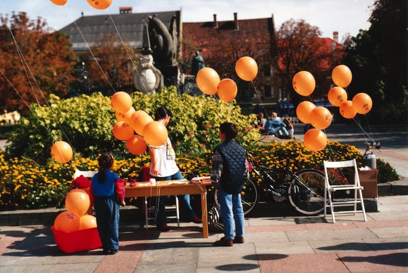 An archival photograph from Independent Curators International's do it archive: a public do it event in a Central European square, with orange balloons printed 'do it'. ICI publishes the photograph without a caption or date, so it cannot be confirmed as the 1994 Klagenfurt exhibition.