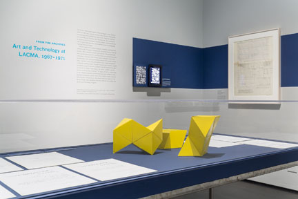 An installation view of the 2015 LACMA exhibition, with the show's blue wall and title panel, framed archival documents, flat display cases and a folded yellow sculptural form on a low plinth.