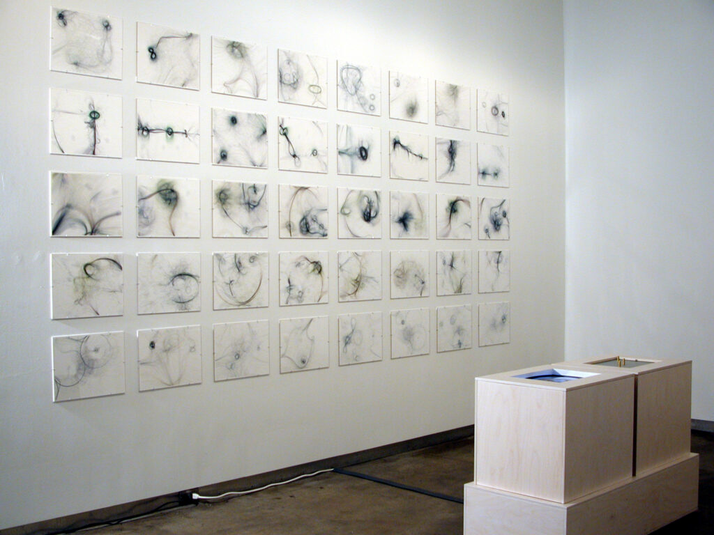 An installation view from The Art of Code at bitforms, New York, with a grid of some forty Tissue prints by Casey Reas on the wall and a plinth-mounted interactive station at right.