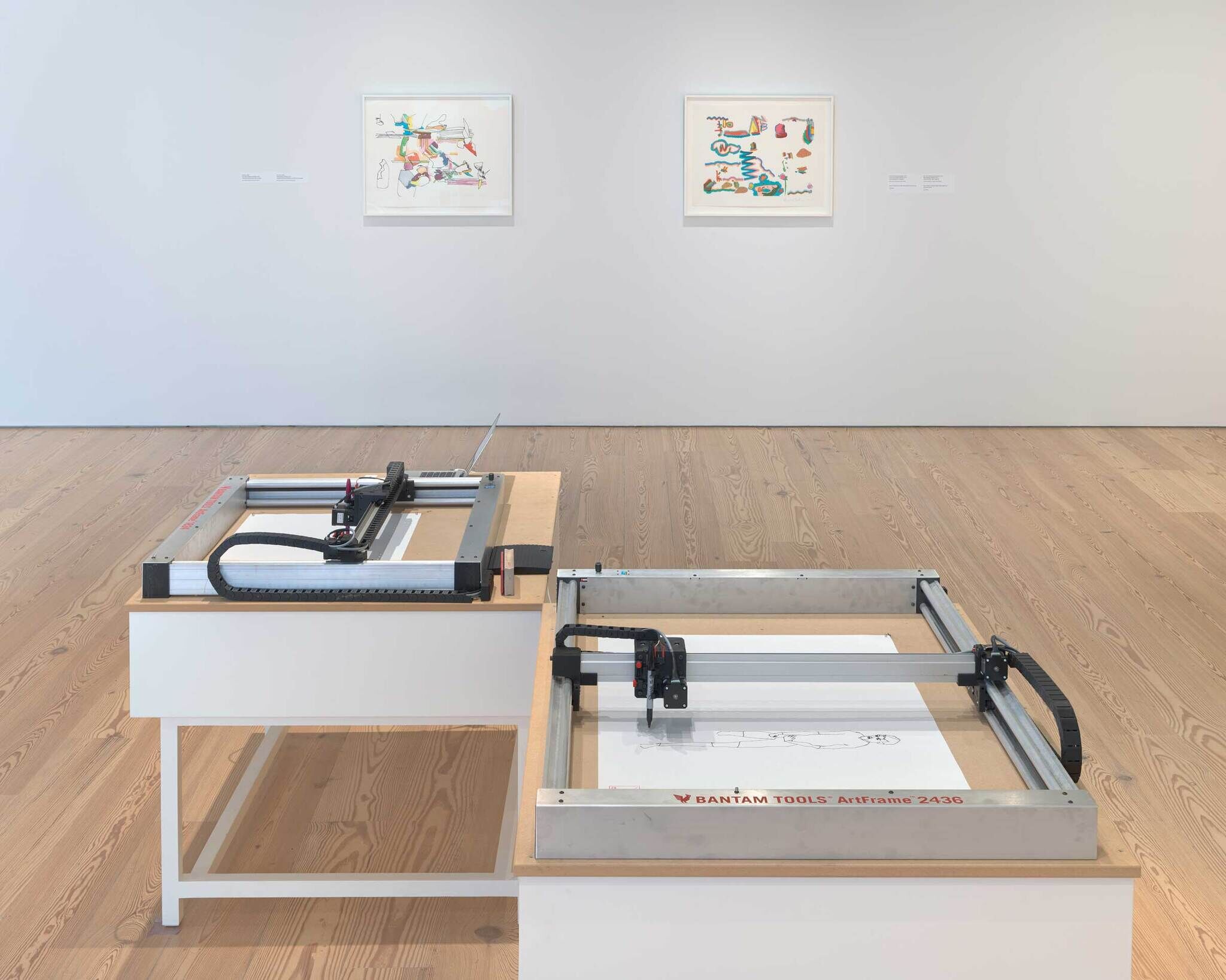 Two active plotters draw AARON images in front of framed Harold Cohen works at the Whitney.