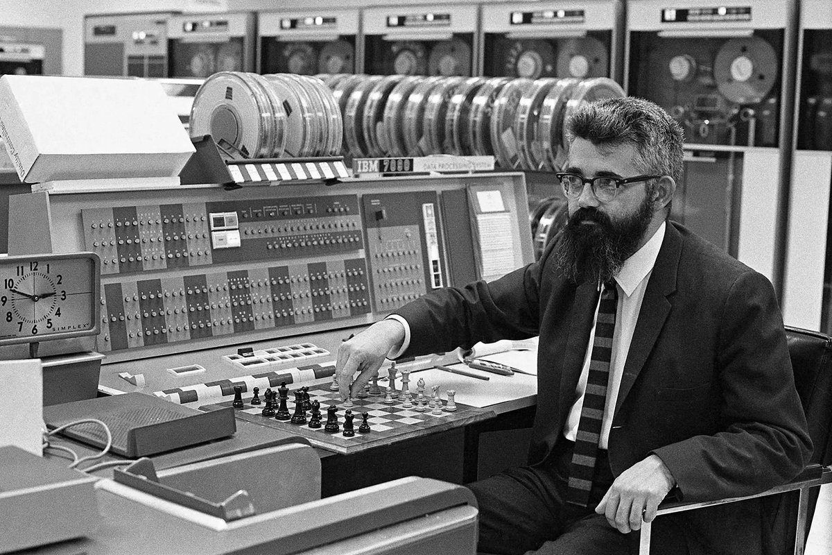 John McCarthy seated at a chessboard beside an IBM 7090 console, with rows of tape reels behind him.