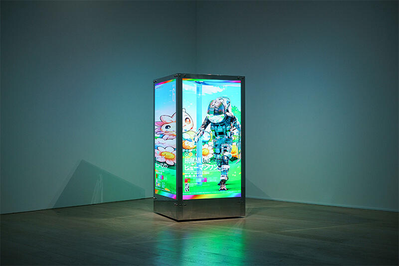 Beeple’s HUMAN ONE video sculpture glows cyan and green in a dark gallery, with colored light reflected across the floor.