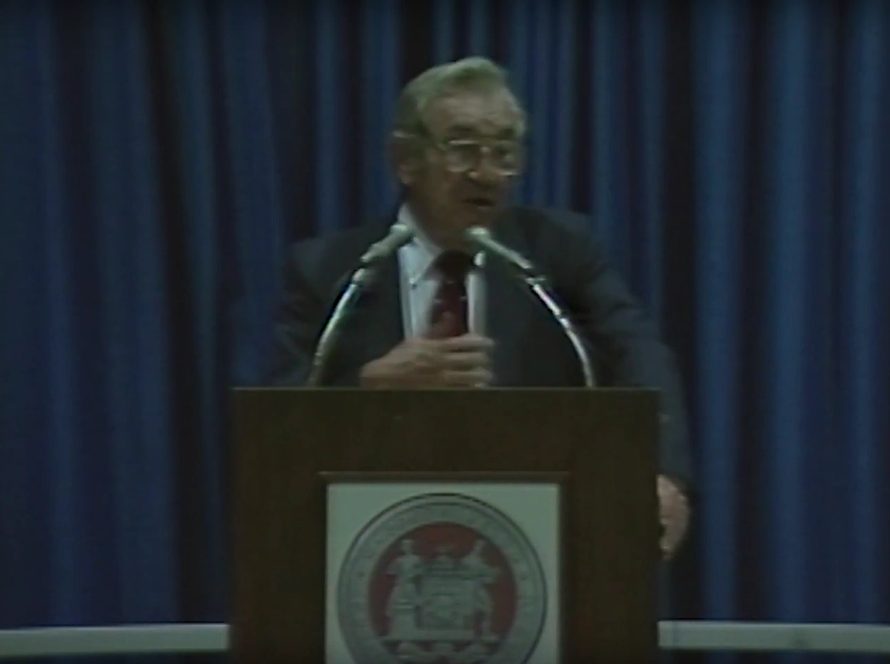 Jerome Wiesner speaks at a lectern with two microphones and an MIT seal, against blue curtains.
