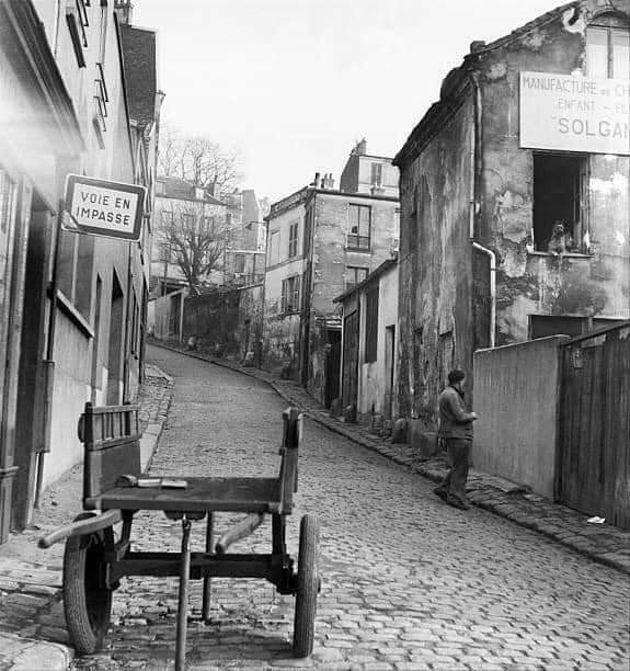 Cobbled Belleville street in 1952 with a handcart, worn buildings, and a pedestrian beside a fence.