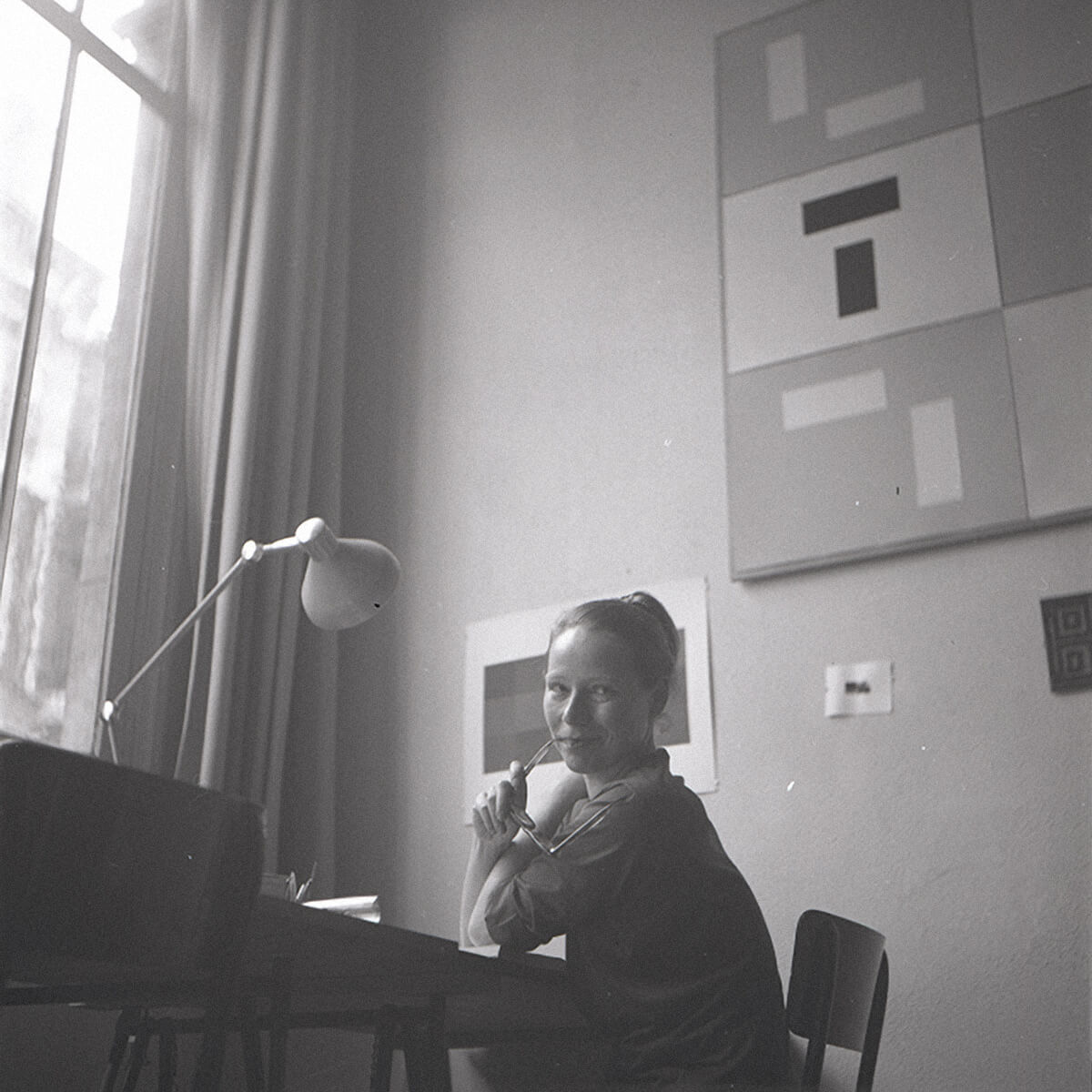 Vera Molnár seated by a tall studio window in 1954, with geometric artworks on the wall behind her.