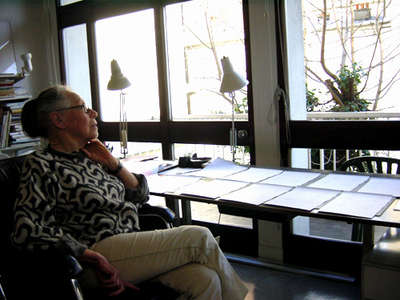Vera Molnár seated in her studio beside a window and a long table of drawings — a much later photograph, filed by Compart only as “Molnar, atelier”.