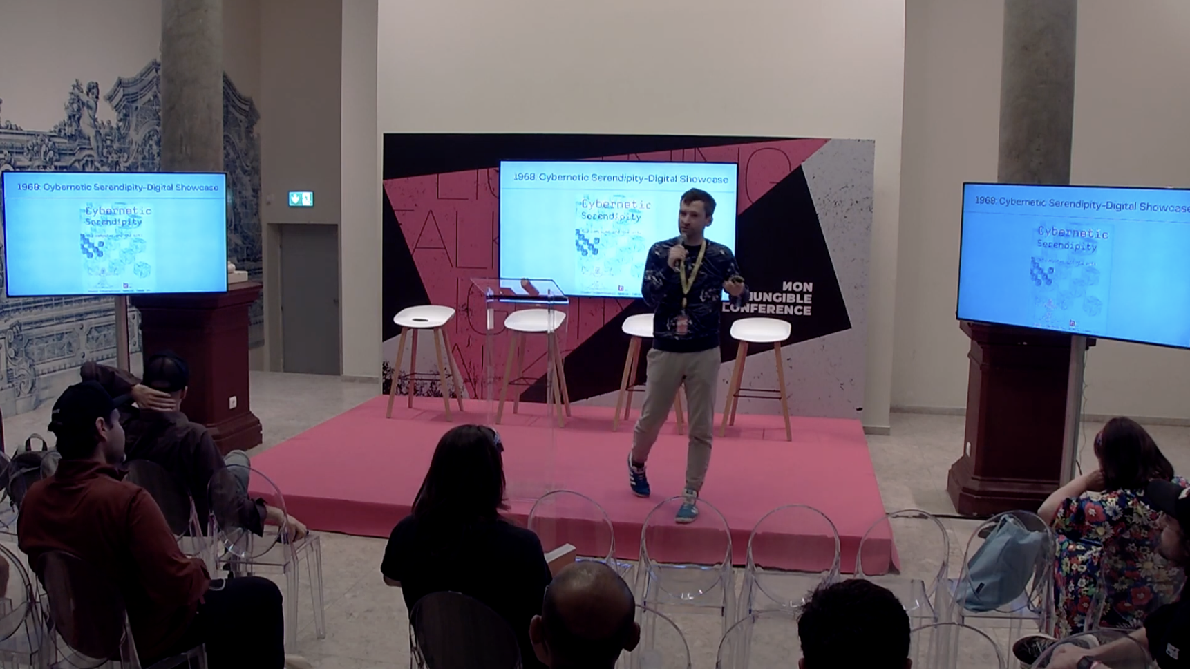 Peter Bauman speaks on a pink stage beside screens showing a diagram of generative art.