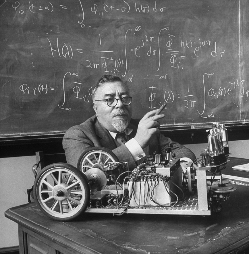 Norbert Wiener holds a cigar beside a wheeled cybernetic moth on a table, with handwritten equations covering the blackboard behind him.
