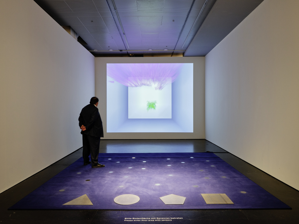 Peter Weibel interacts with an installation comprising a purple carpet marked with sensor dots and geometric shapes beneath a projection of a recursive virtual room.