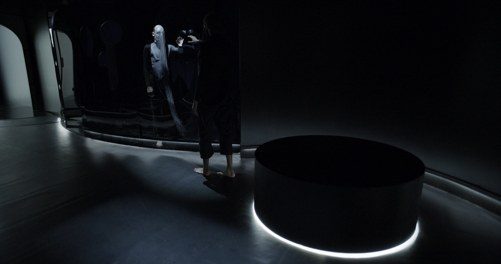 Installation view of Operator's On View at the SCAD Museum of Art: a darkened gallery with a lit circular plinth and a figure reflected in a curved mirrored wall.