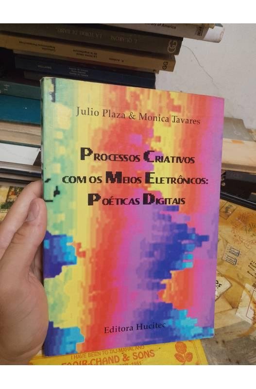 A bookseller's photograph of a copy of Processos criativos com os meios eletronicos: poeticas digitais held in the hand, showing the pixelated rainbow cover with the authors' names and the Hucitec imprint.