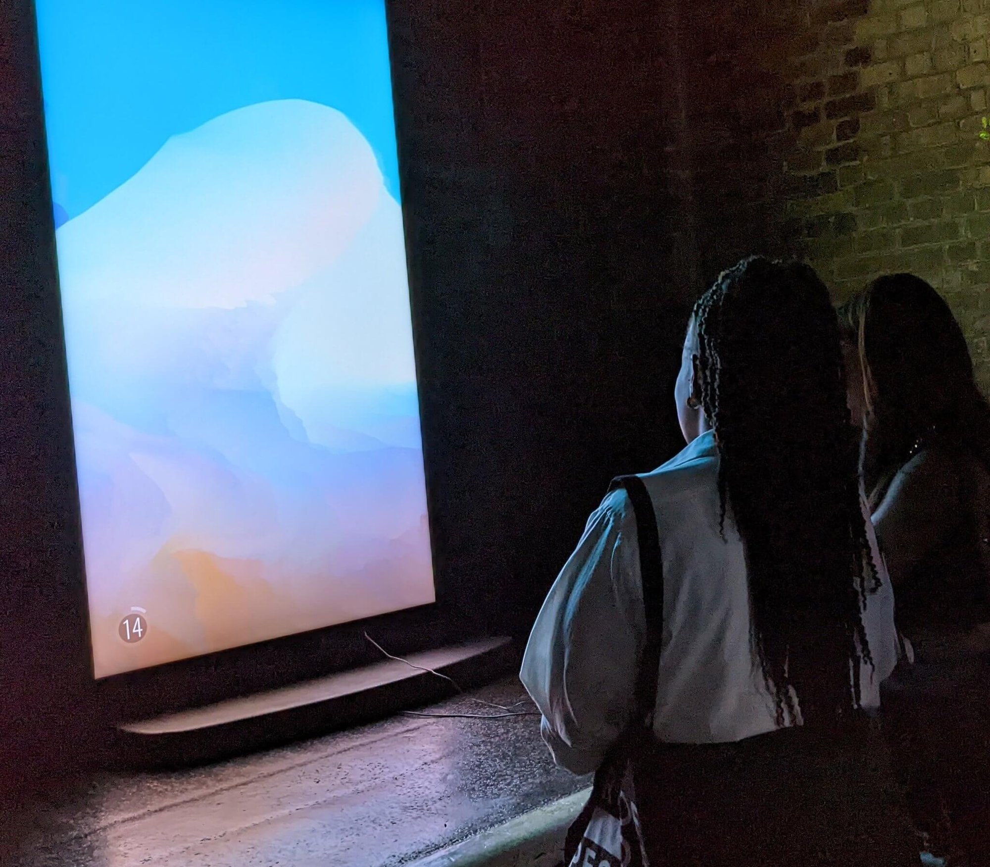 Two visitors stand in a brick-walled space before the luminous cyan, pink and lavender screen of Mario Klingemann’s Dy5p1ace.