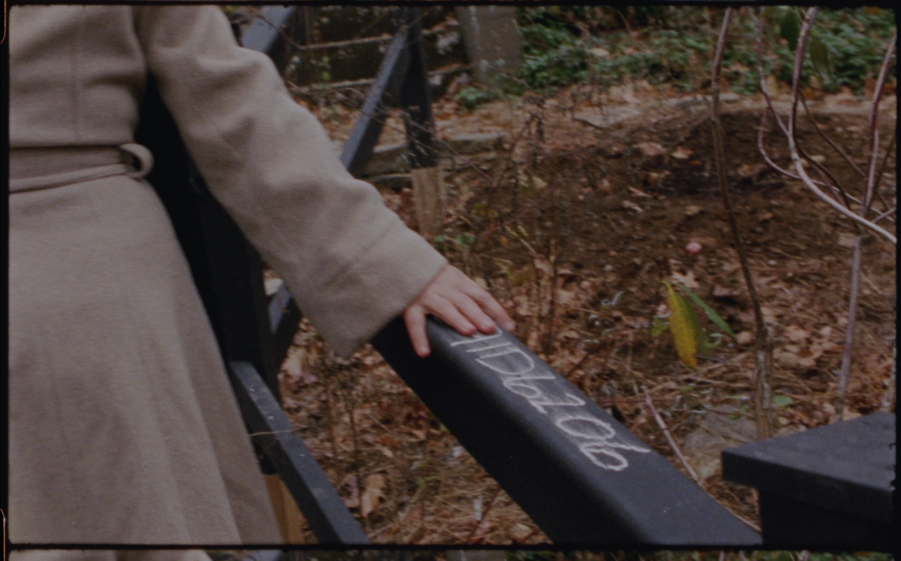 A woman in a light coat rests her hand on a dark outdoor stair railing marked with a sequence of handwritten characters.