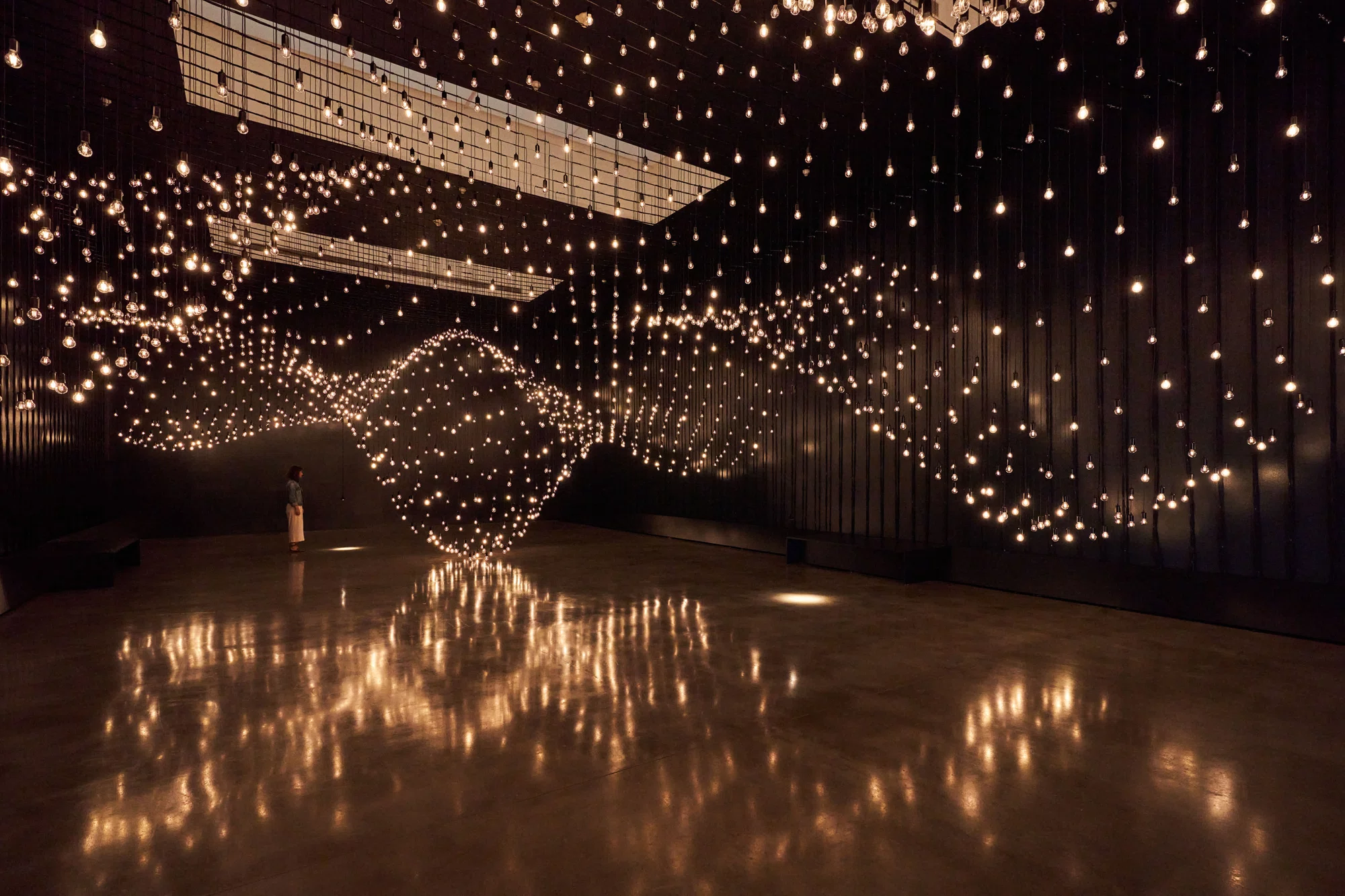 An installation view of the exhibition, not a single work in isolation: Lozano-Hemmer's Pulse Topology filling a blacked-out gallery with thousands of suspended dimmable bulbs pulsing at visitors' heart rates, their light pooling on the polished floor.