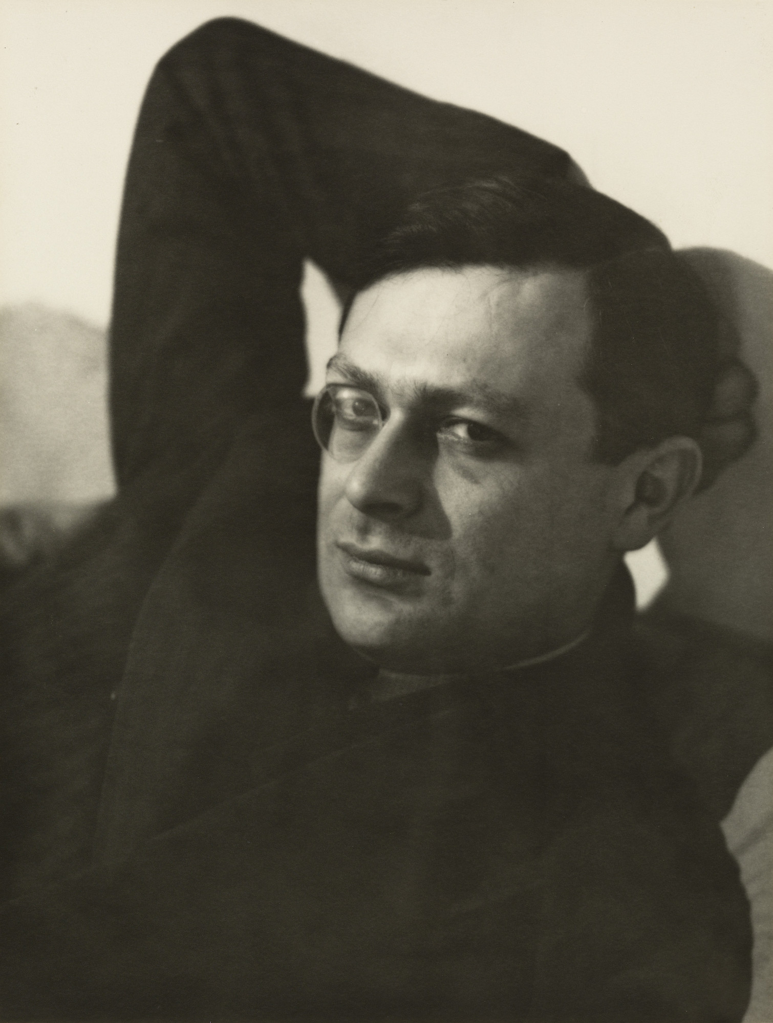 Tristan Tzara wearing a monocle, photographed with his arms raised behind his head and a strong shadow on the wall.