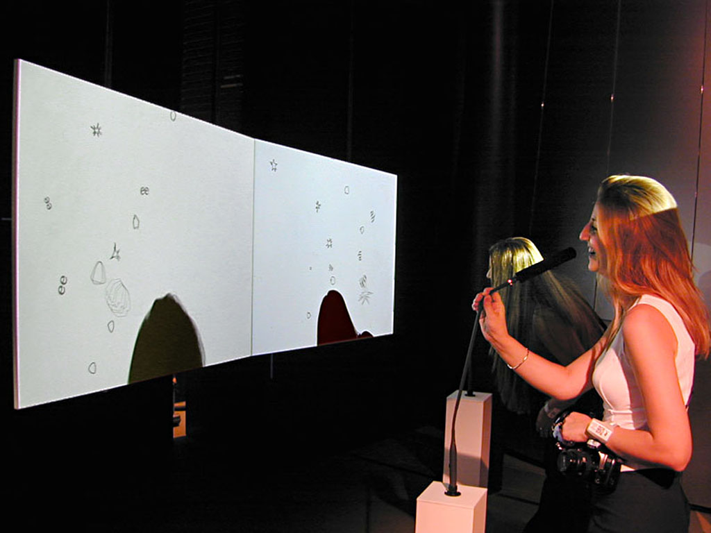 An installation view of Re:MARK: a visitor speaks into a microphone while her silhouette on the projection screen emits animated glyphs and shapes generated from her voice.