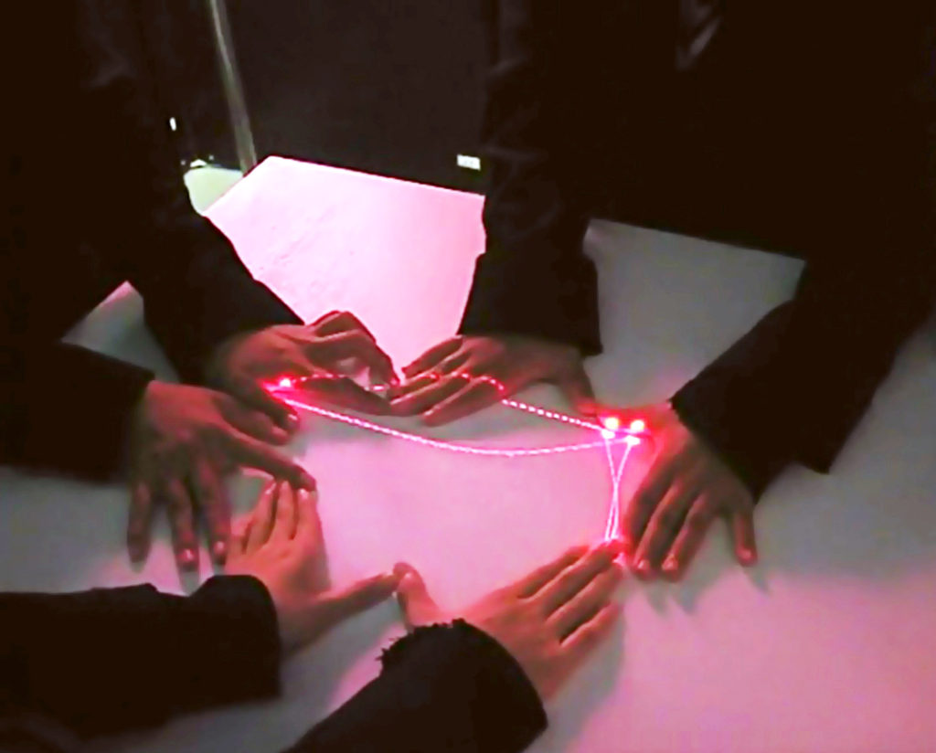 scoreLight in use: several pairs of hands around a sheet of paper as the instrument's red laser spot traces the drawn contour it is reading as sound.