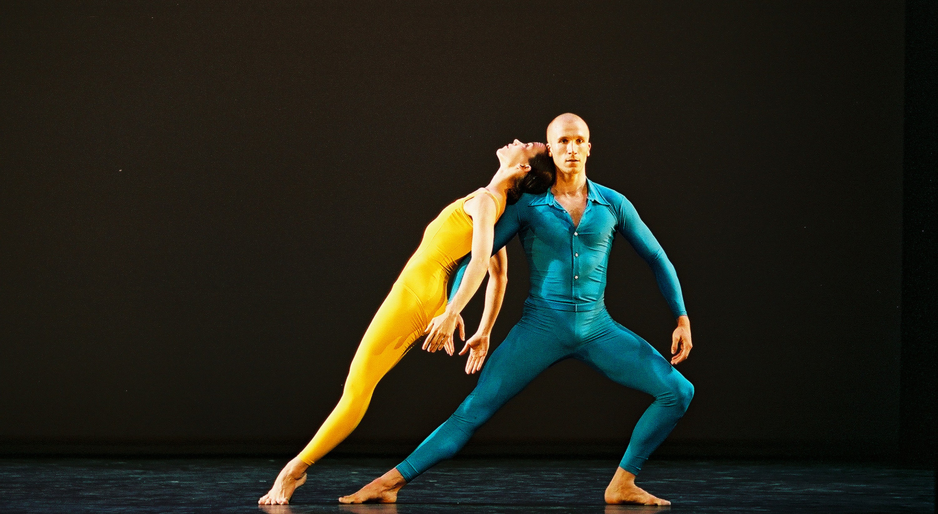 Two dancers in yellow and blue leotards extend their legs and arms against a black stage.
