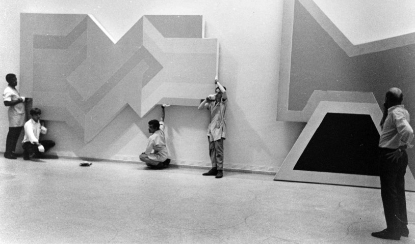A team holds a large angular geometric painting against a gallery wall while Lawrence Alloway stands at right beside another shaped canvas.