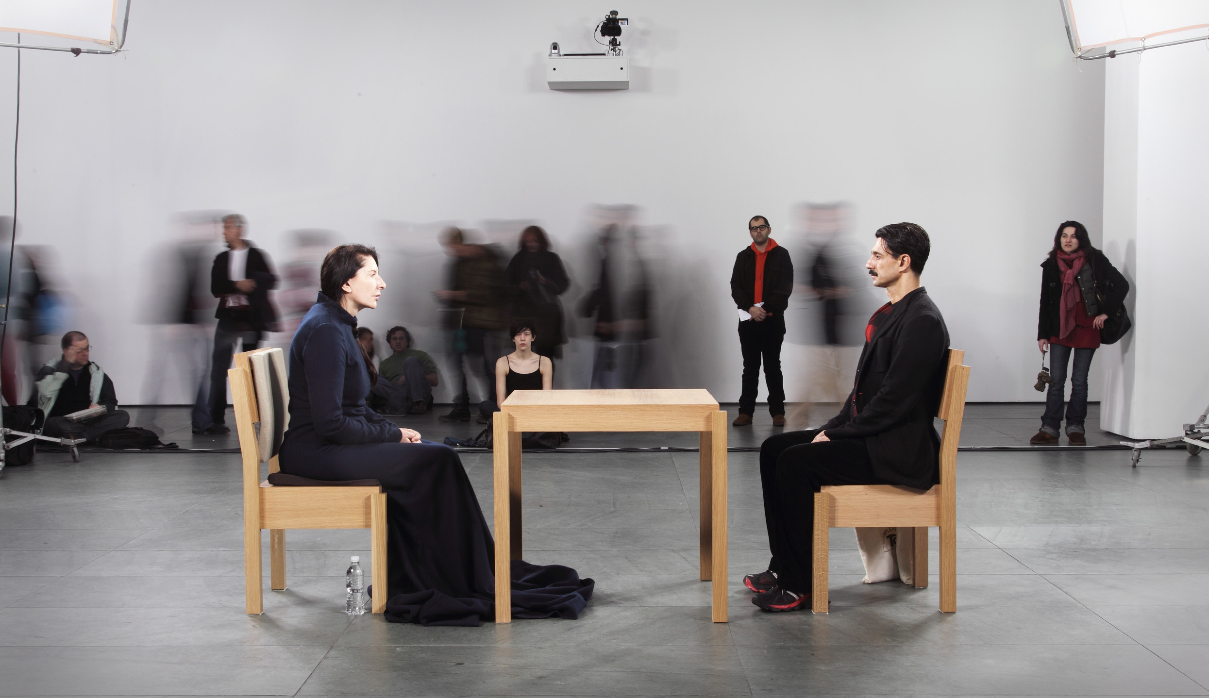 Abramović and a seated participant face one another across a table while museum visitors stand behind them.