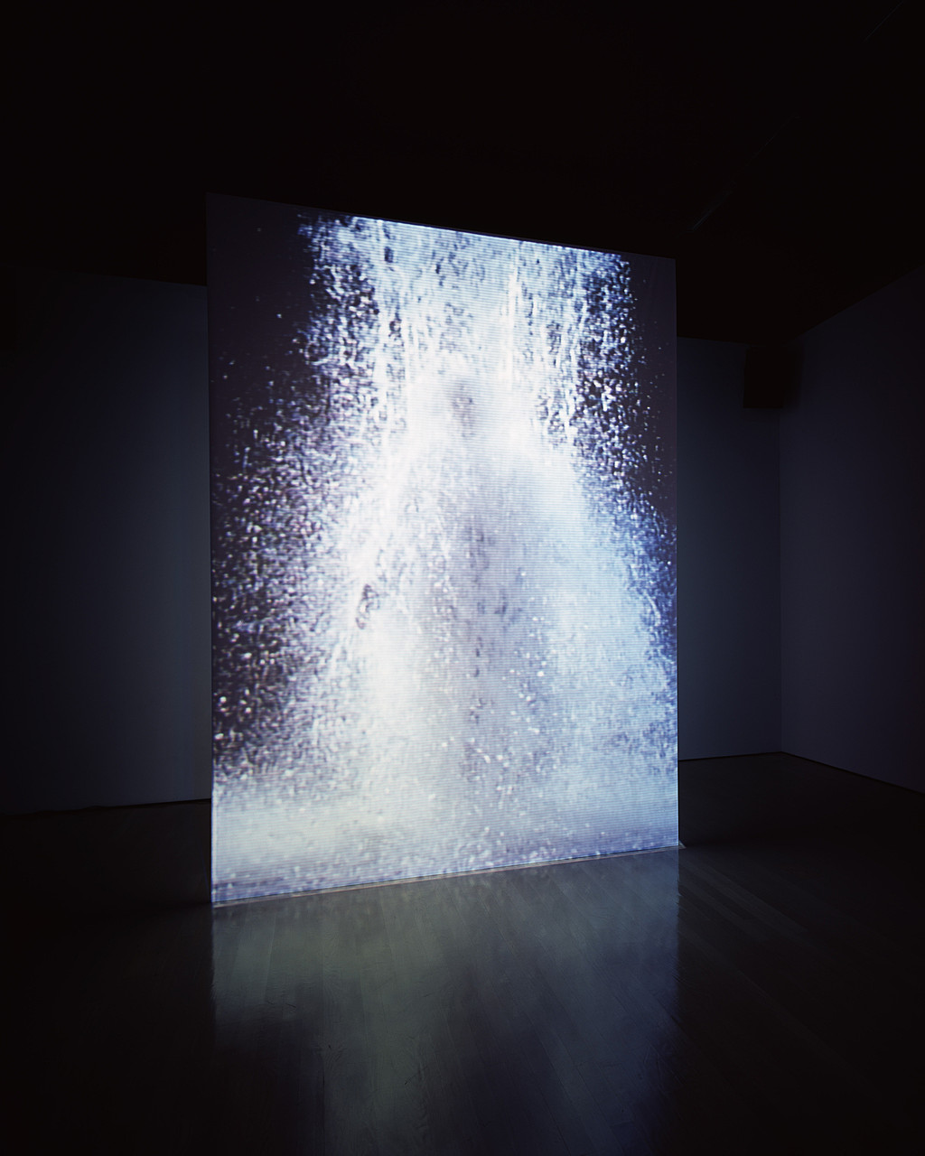 An installation view of Bill Viola's The Crossing, showing the water channel cascading over the figure on one face of the free-standing double-sided screen.