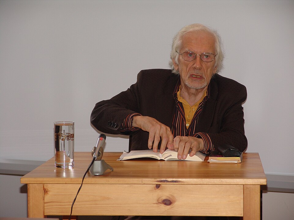 A photographic portrait of Herbert W. Franke, author of 'The Expanding Medium: The Future of Computer Art', reading from a book at a table with a microphone in 2007; it is a portrait of the author, not an image of the essay.