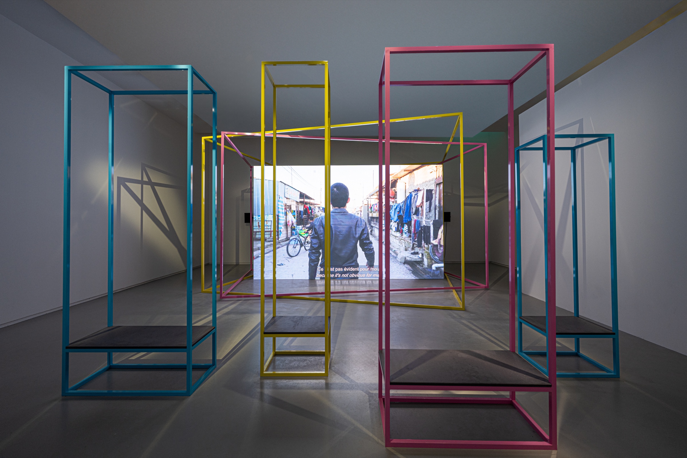 Pink, yellow and cyan open frames create a layered geometric space in front of the video screen in Hito Steyerl’s Mechanical Kurds installation.