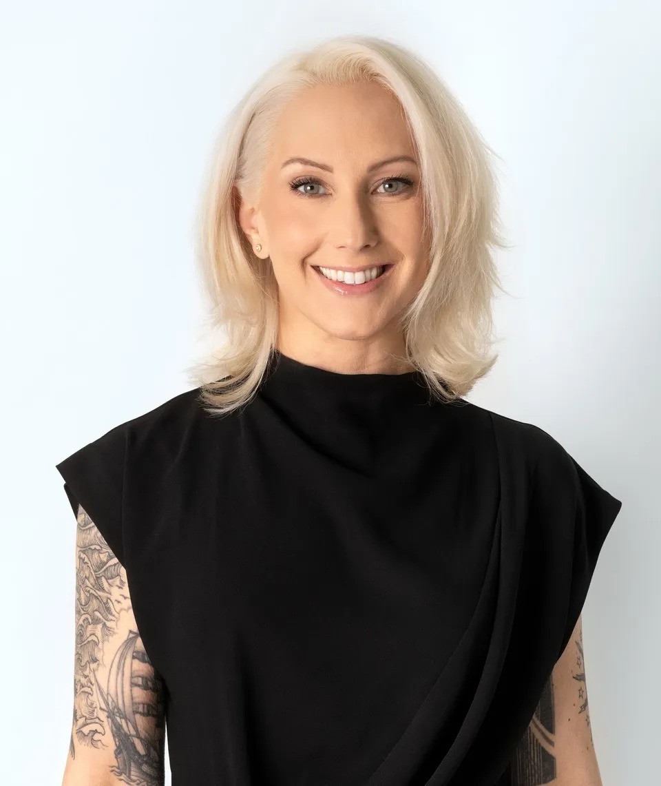 Tina Rivers Ryan smiles at the camera, wearing a sculptural black top against a pale background.