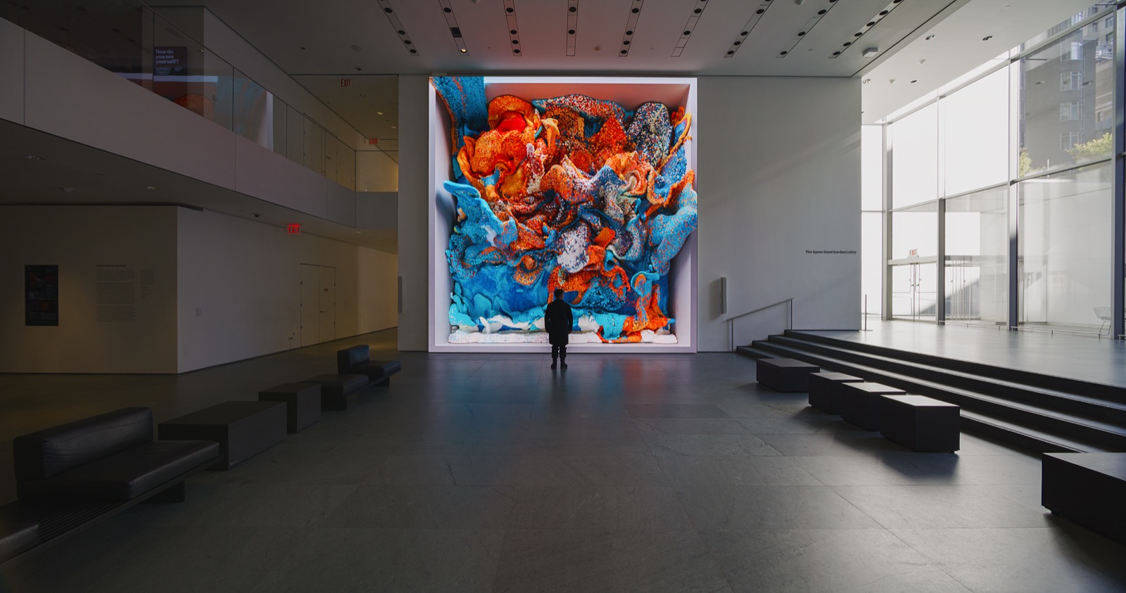 Refik Anadol's Unsupervised installed at the Museum of Modern Art: a single tall LED wall in the museum's ground-floor lobby churning through a mass of orange and blue machine-generated form, with one visitor standing in silhouette before it and the lobby's benches and steps to either side.