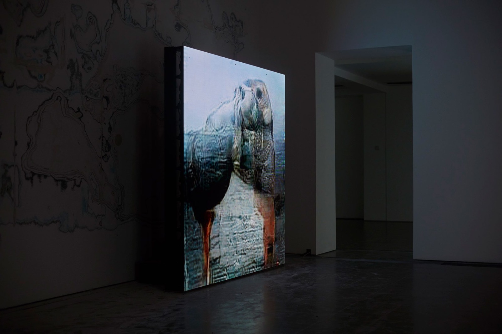 Installation view of UUmwelt at the Serpentine: one of the large freestanding LED screens showing a neural-network reconstruction, in a gallery whose walls carry the work's insect-marked surfaces.