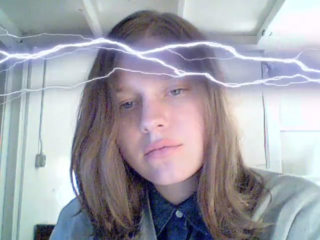 A still from Petra Cortright's VVEBCAM: the artist looking impassively into her laptop webcam while animated lightning-bolt clip art streaks across her forehead.