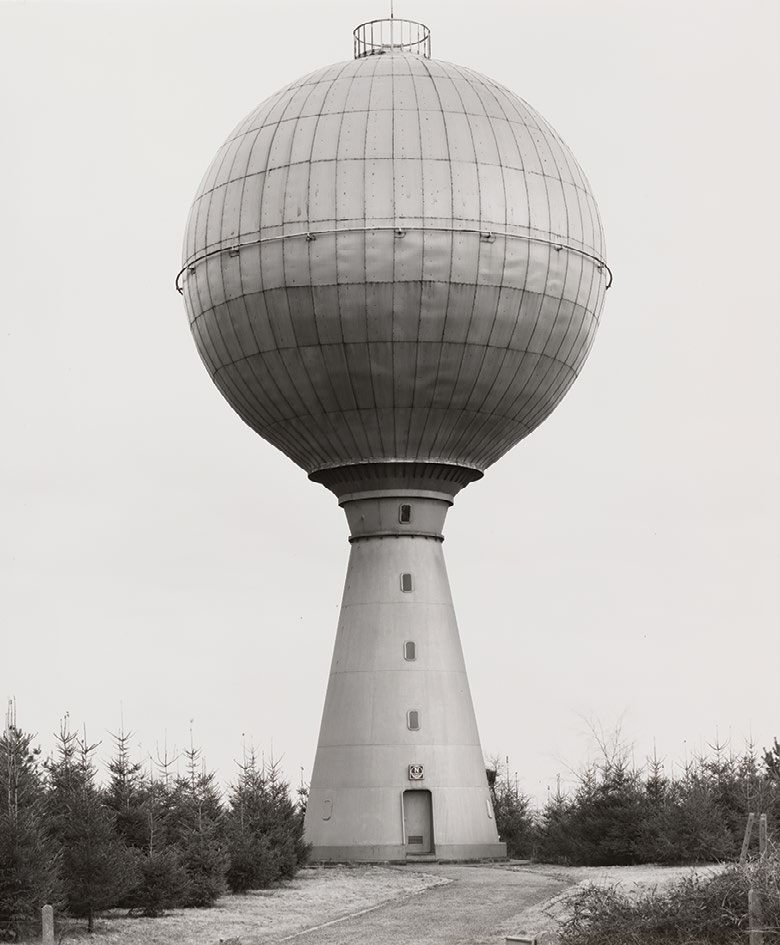 Black-and-white photograph of a spherical water tank raised on a tapered concrete tower, framed by low trees and an overcast sky.