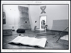 Black-and-white installation view showing sculptures and interventions across a gallery floor and walls.