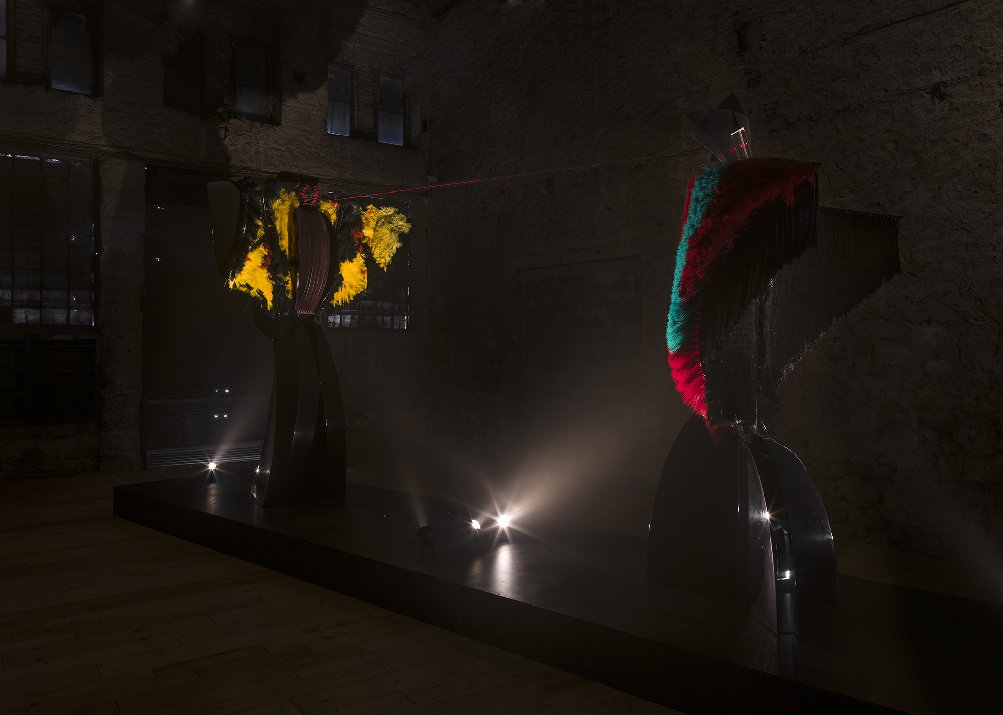 Liliane Lijn’s two winged kinetic sculptures face one another in a dark gallery, their prism heads connected by a red laser beam.