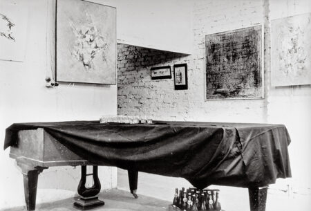 Black-and-white view of paintings hanging on the walls of a brick studio, with a cloth-covered piano in the foreground.